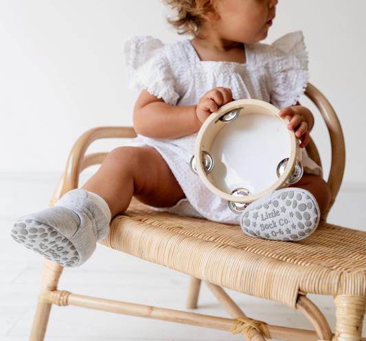Child sitting on a wicker chair holding a tambourine with 'Little Sock Co.' branding. STAY-ON, NON-SLIP BOOTIE - GREY | THE LITTLE SOCK CO.