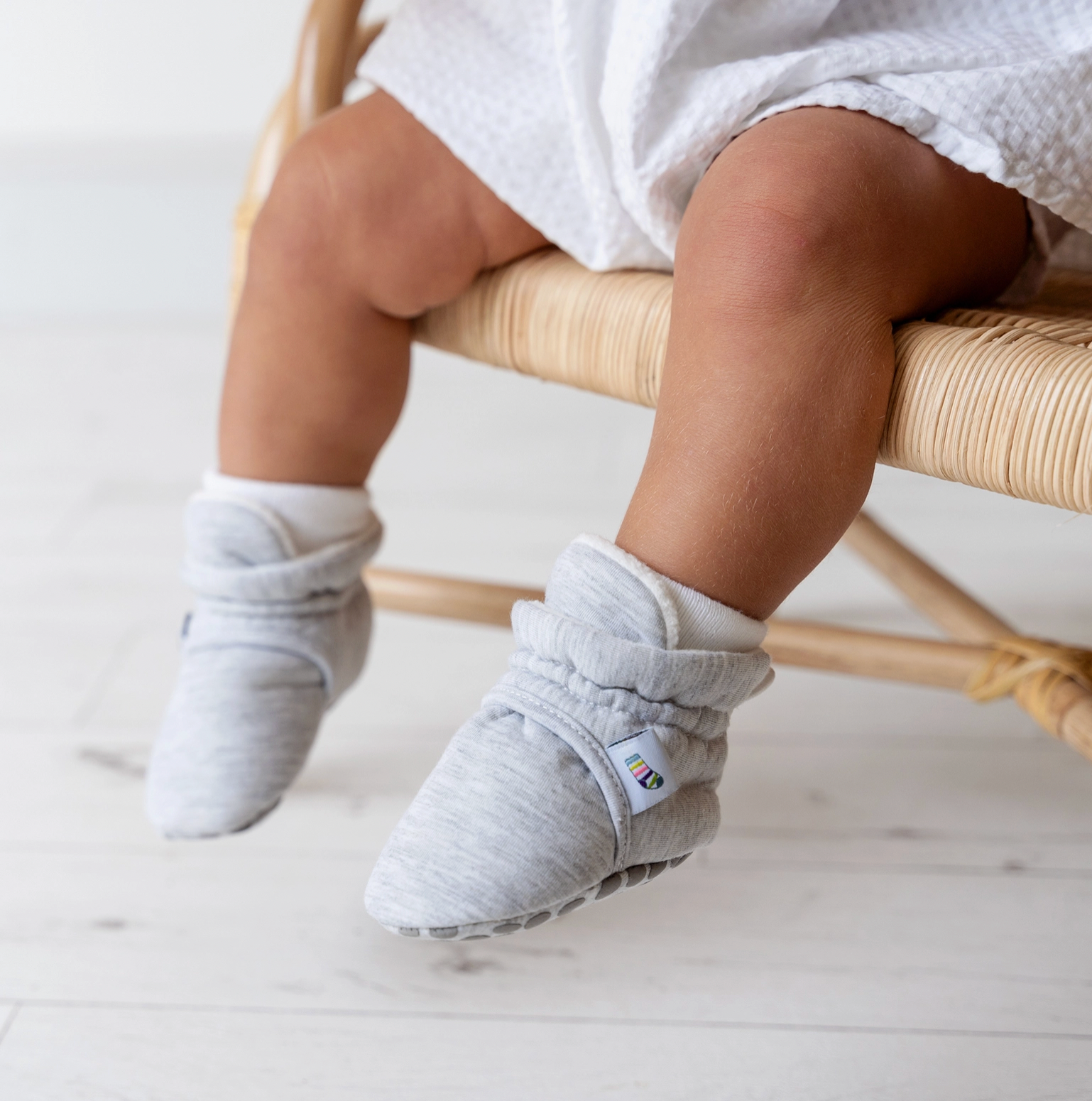 Baby's feet wearing gray shoes with a visible brand logo, sitting on a light wooden stool. STAY-ON, NON-SLIP BOOTIE - GREY | THE LITTLE SOCK CO.