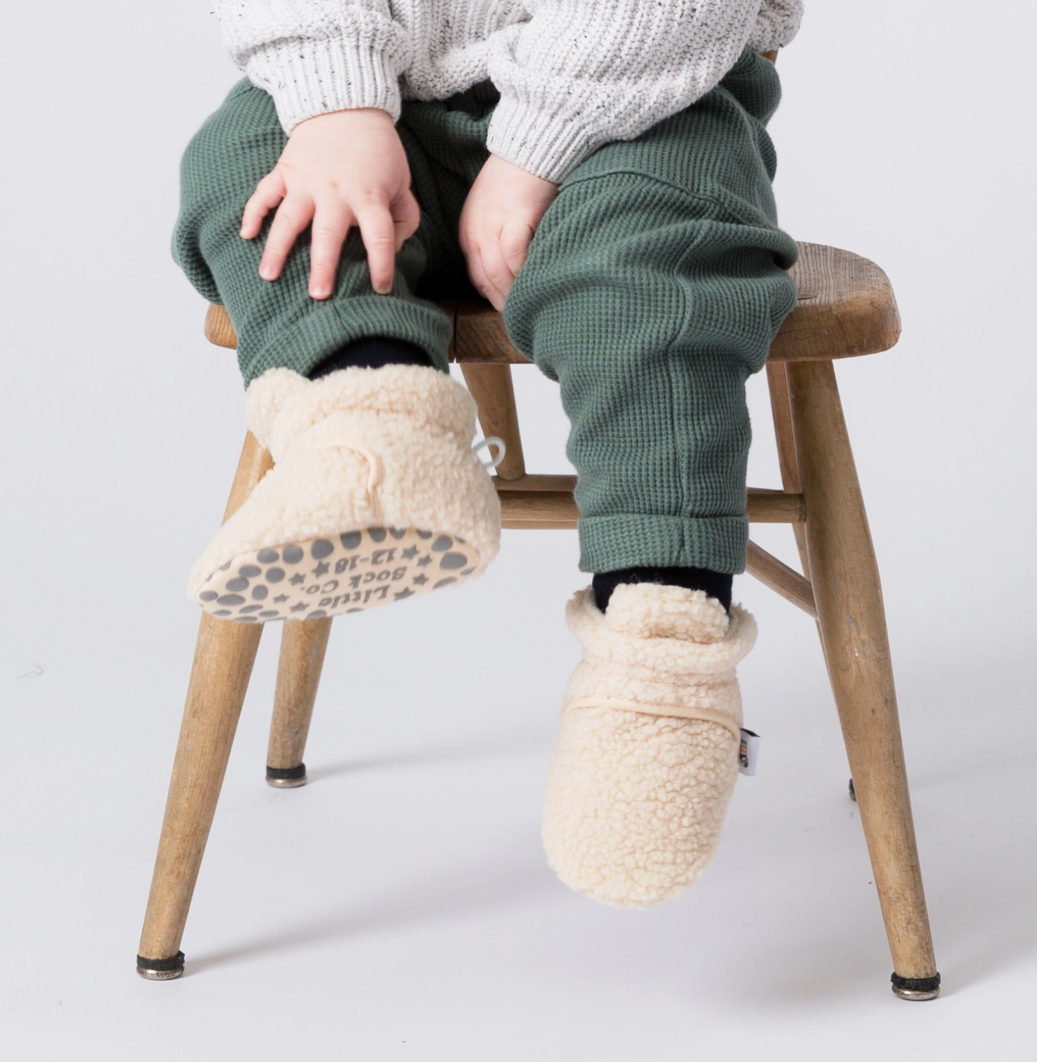 Person wearing white fuzzy slippers sitting on a wooden stool against a light background STAY-ON, NON-SLIP BOOTIE - SHERPA | THE LITTLE SOCK CO.