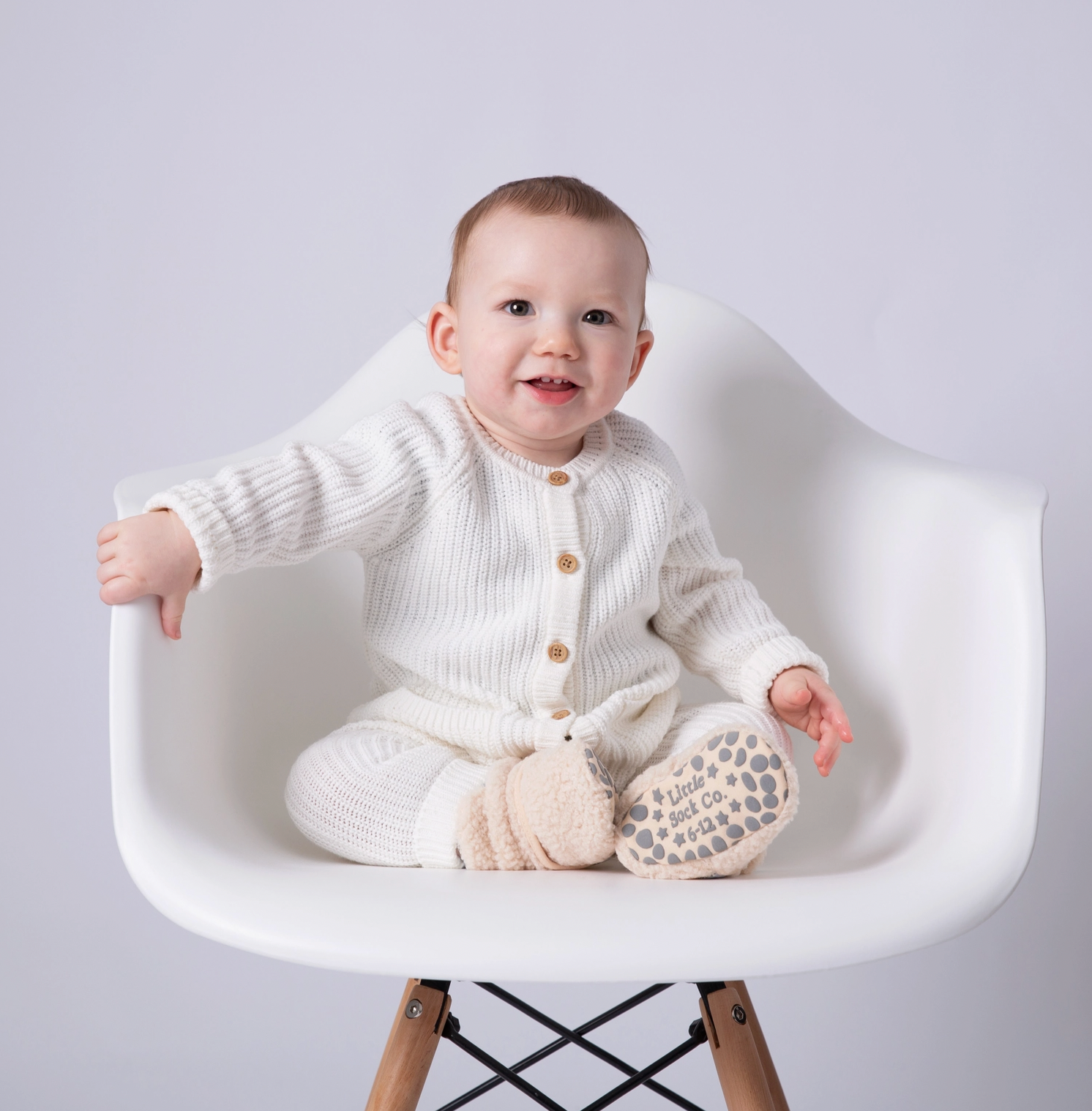 Baby in a white knit outfit sitting on a white chair with a plain background STAY-ON, NON-SLIP BOOTIE - SHERPA | THE LITTLE SOCK CO.