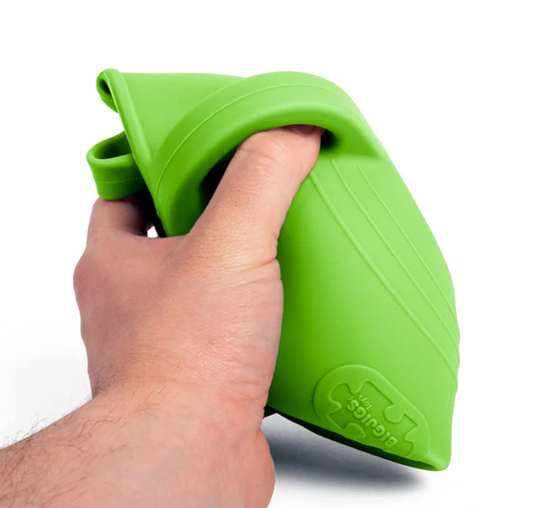 Bigjigs lime green silicone children's watering can — adult hand squeezing and squashing the can to demonstrate flexible, shatterproof silicone material, with embossed Bigjigs Toys logo visible, on white background