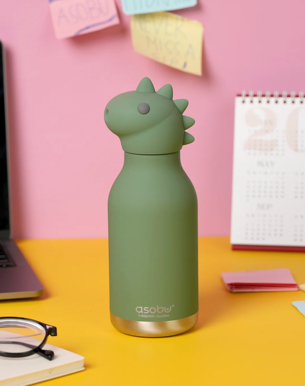 A matte sage green Asobi dinosaur-shaped water bottle with a gold-toned base, standing on a yellow desk against a pink wall with sticky notes and a calendar.