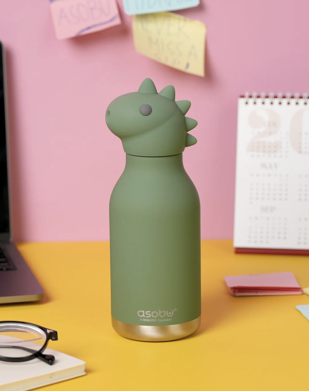 A matte sage green Asobi dinosaur-shaped water bottle with a gold-toned base, standing on a yellow desk against a pink wall with sticky notes and a calendar.