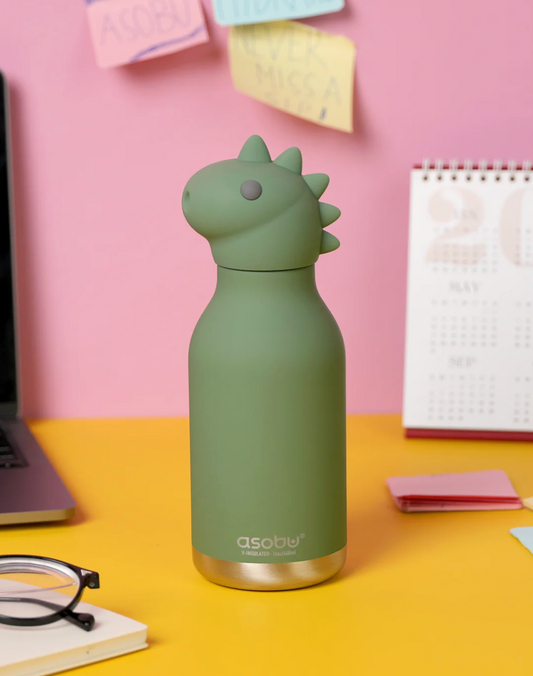 A matte sage green Asobi dinosaur-shaped water bottle with a gold-toned base, standing on a yellow desk against a pink wall with sticky notes and a calendar.