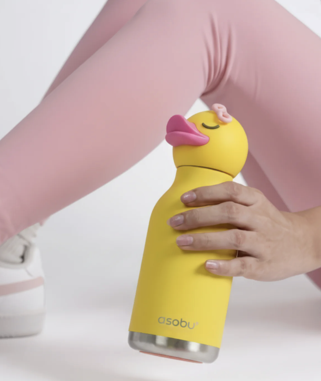 A person in pink leggings holds a yellow duck-shaped water bottle with a pink lid and a stainless steel base. The brand name 'asobu' is printed near the bottom of the bottle.