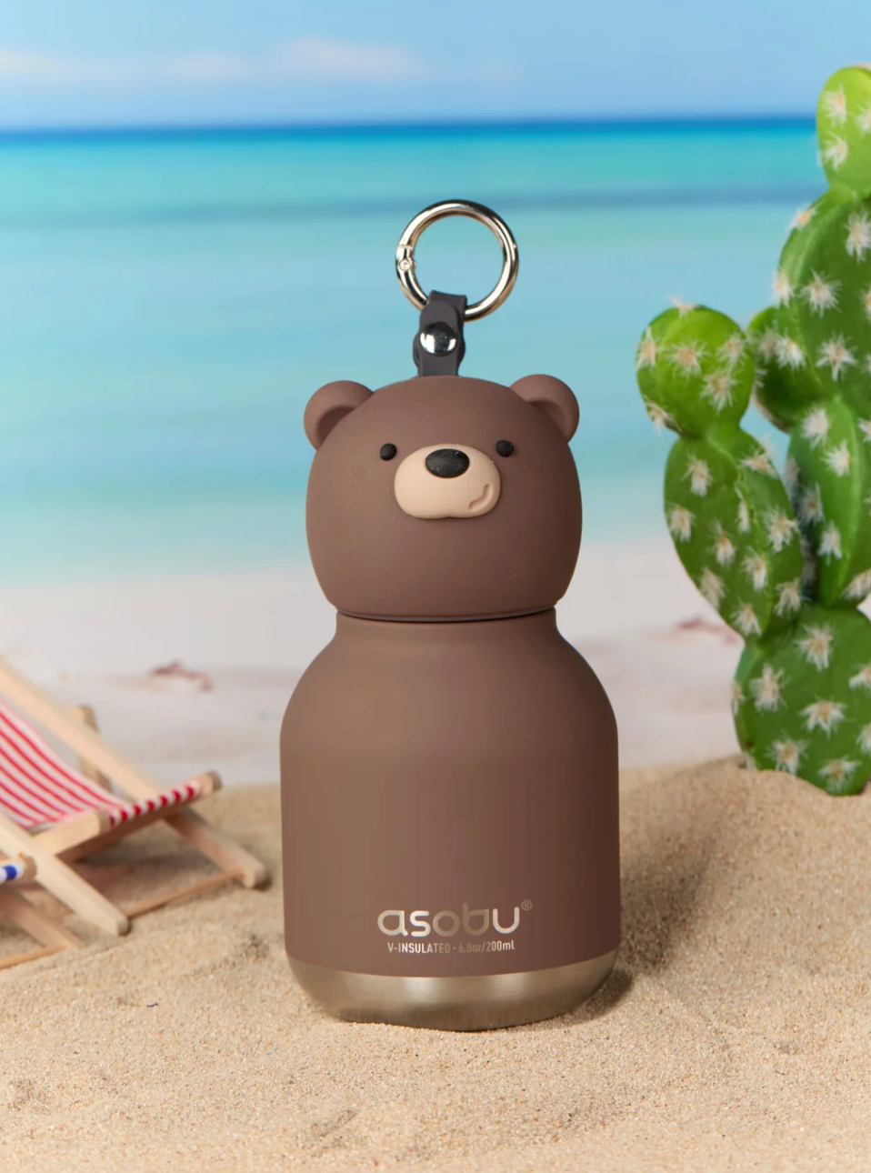 A brown bear-shaped insulated bottle with a metal key ring, standing in sand with a beach scene and cactus in the background.