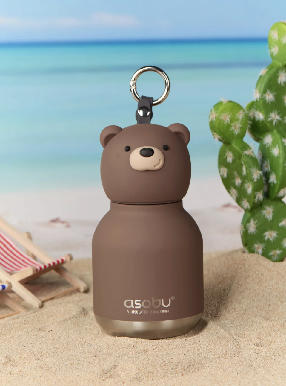 A brown bear-shaped insulated bottle with a metal key ring, standing in sand with a beach scene and cactus in the background.