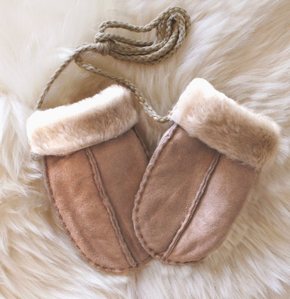 A pair of beige suede moccasin-style slippers with cream faux-fur trim and a braided rope handle, laid flat on a white faux-fur surface.