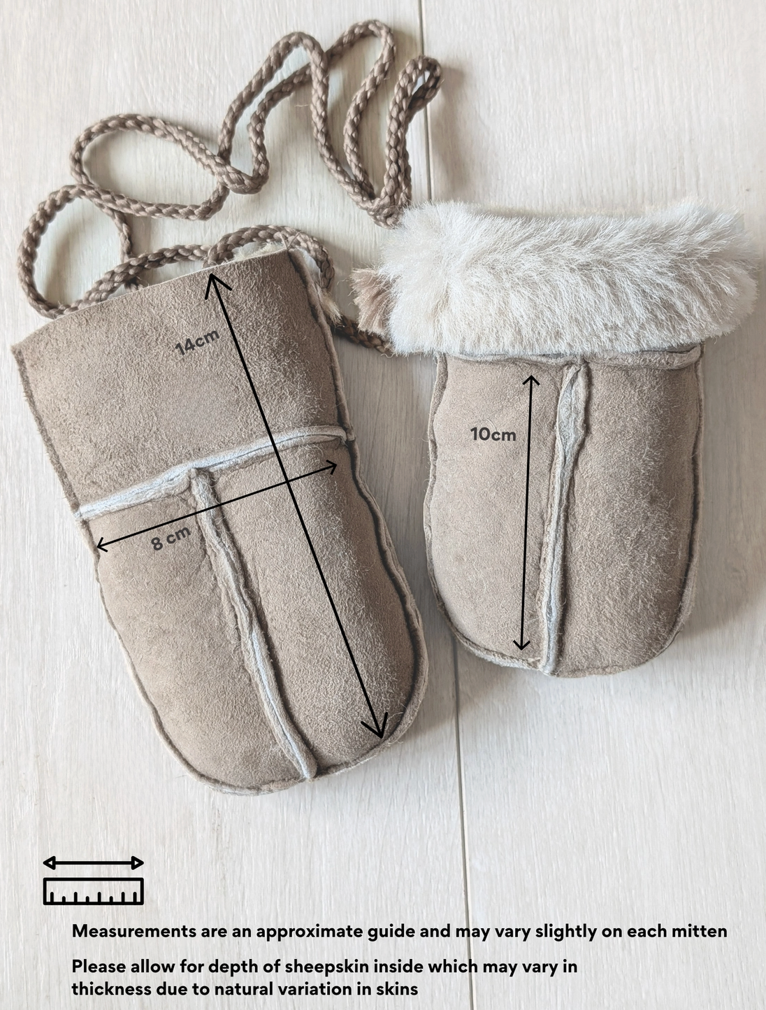 Two beige suede-like baby mittens laid flat on a light wood background, one with a braided strap and one with faux fur trim. Black measurement arrows and text indicate approximate dimensions of each mitten.