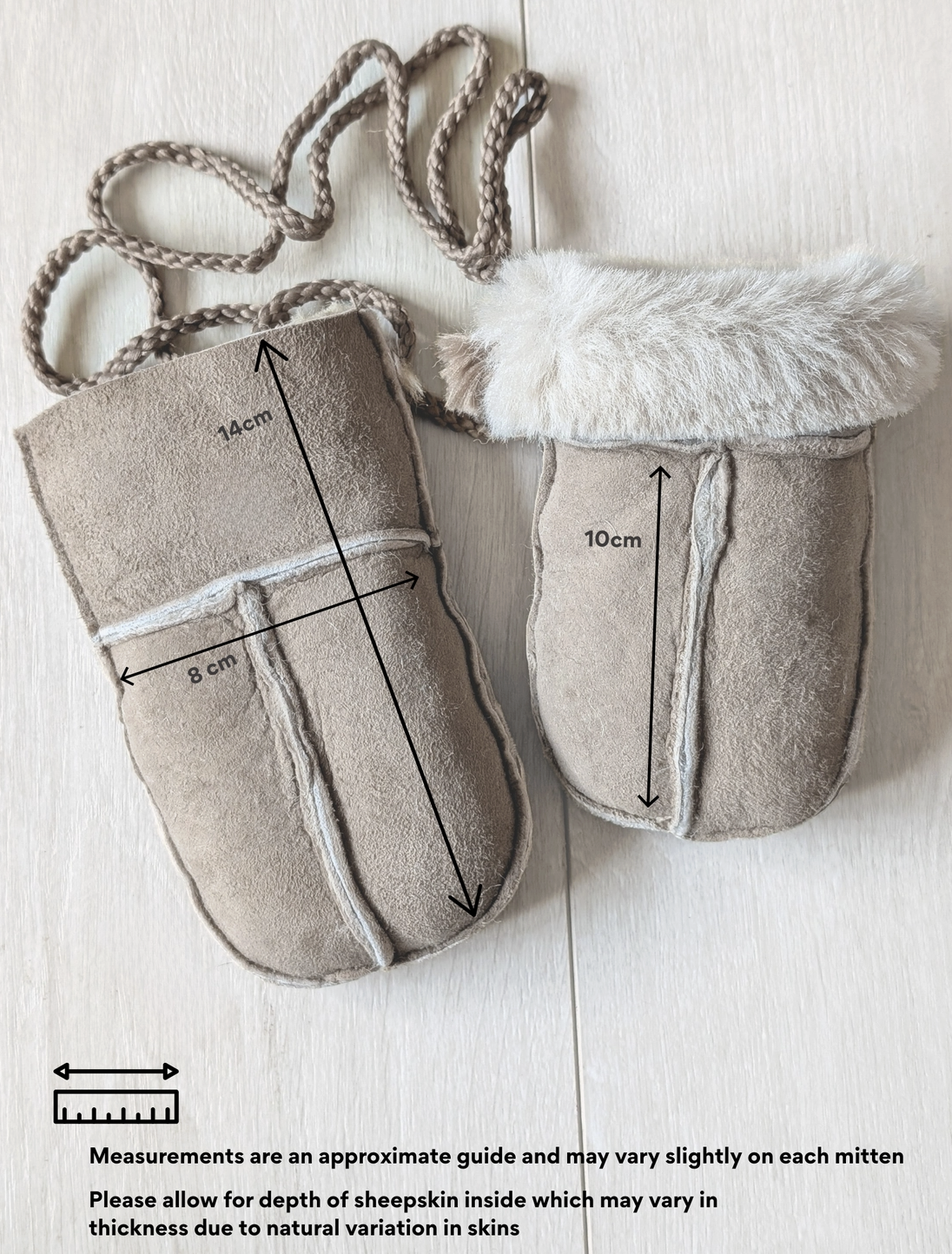 Two beige suede-like baby mittens laid flat on a light wood background, one with a braided strap and one with faux fur trim. Black measurement arrows and text indicate approximate dimensions of each mitten.