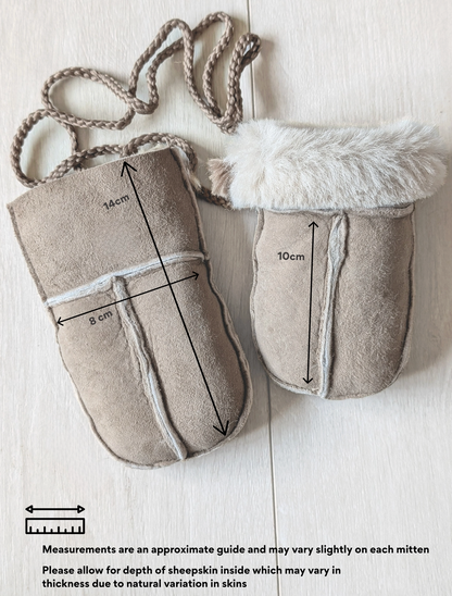 Two beige suede-like baby mittens laid flat on a light wood background, one with a braided strap and one with faux fur trim. Black measurement arrows and text indicate approximate dimensions of each mitten.