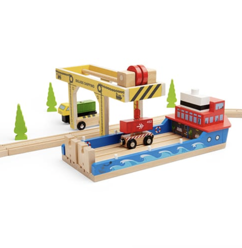 A wooden toy port and cargo train set on a white background, featuring a blue boat with a red cabin, a yellow crane, a small green truck, and green trees.