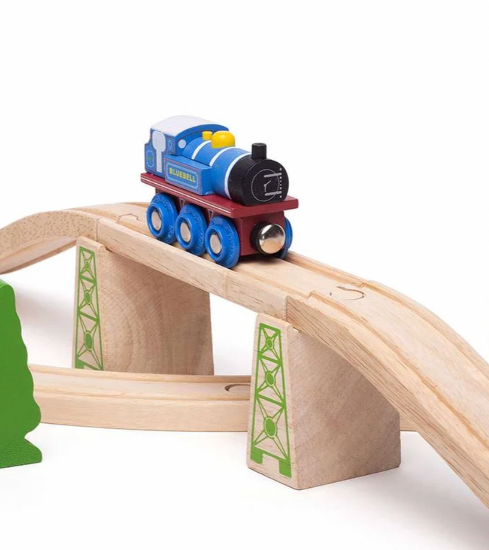 A wooden toy train with a blue body, black front, and red undercarriage, traveling on a light natural wood bridge with green-trimmed supports against a white background.