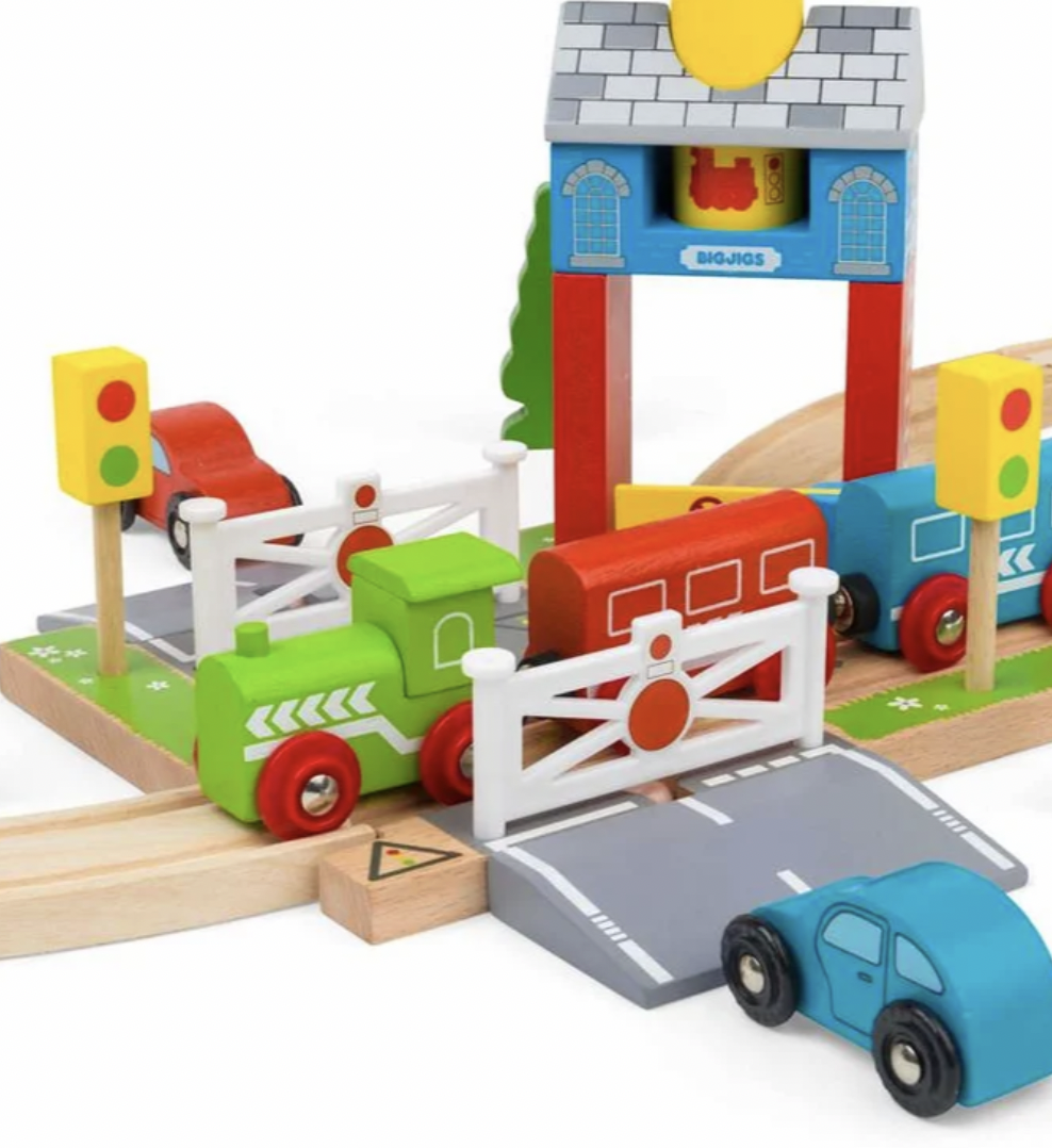 A colorful wooden toy train and car set on a white background, featuring a green train, blue and red cars, a white fence, a gray road, and a blue station building.