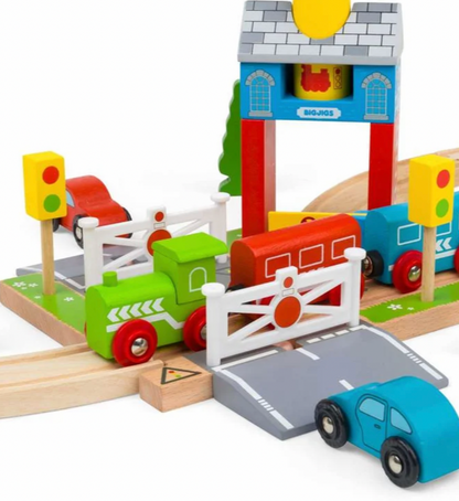A colorful wooden toy train and car set on a white background, featuring a green train, blue and red cars, a white fence, a gray road, and a blue station building.