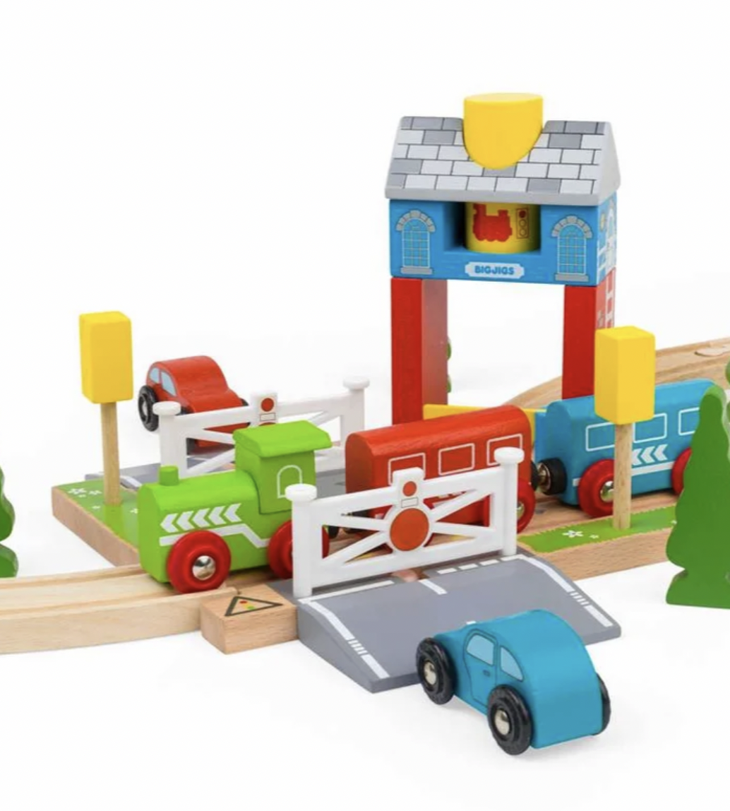 A colorful wooden toy train set on a white background, featuring a green train, blue passenger train, red cargo car, wooden tracks, a station platform with a blue and red signal tower, a white gate, a small blue car, and small yellow trees.