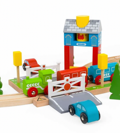 A colorful wooden toy train set on a white background, featuring a green train, blue passenger train, red cargo car, wooden tracks, a station platform with a blue and red signal tower, a white gate, a small blue car, and small yellow trees.
