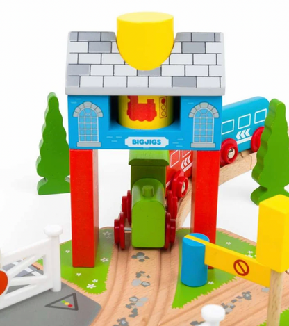 A colorful wooden toy train station set with a blue building, red pillars, a gray roof, green trees, brown tracks, and toy trains in green, red, and blue. The brand name 'BIGJIGS' is visible on the front of the station.