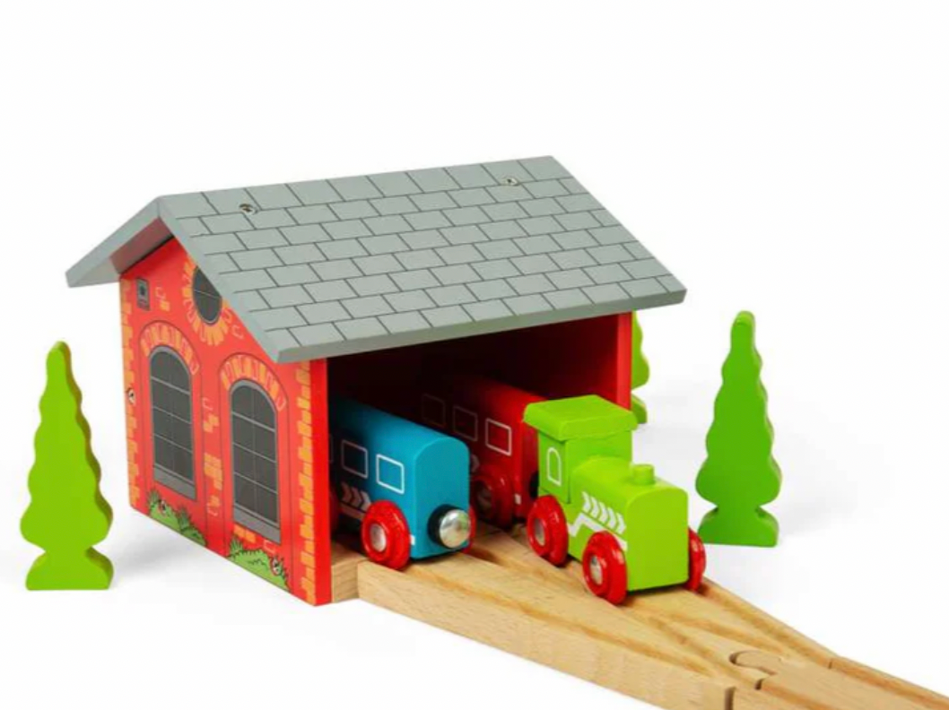 A wooden toy train station set with a red-orange station building, gray tiled roof, light wood tracks, a blue train car, a green train engine, and green trees on a white background.