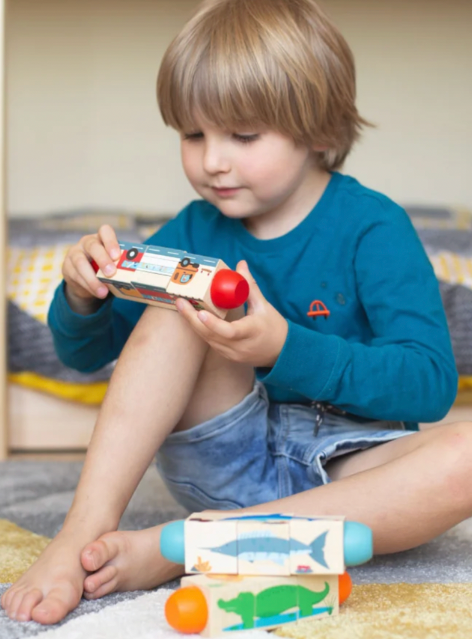 A young child with blonde hair sits on a carpeted floor, holding a small toy block with a red ball on top. The child is wearing a blue long-sleeve shirt and denim shorts.