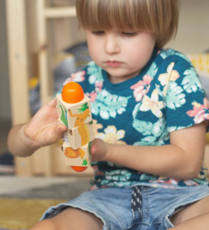 A young child with blonde hair wearing a blue floral t-shirt and denim shorts holds a wooden toy with animal illustrations and orange ends while sitting indoors.