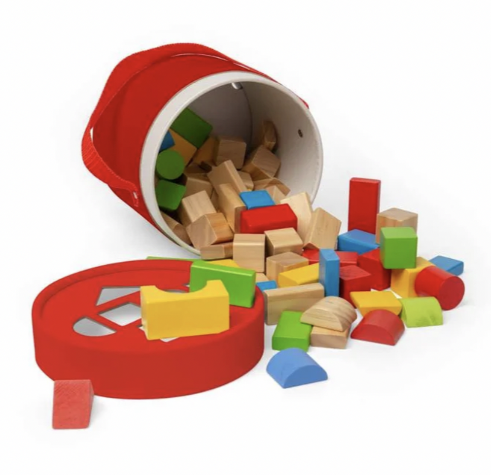 A red children's wooden block storage bin tipped over with colorful wooden blocks spilling out onto a white background. The set includes a red bucket-shaped container, a round lid, and multicolored blocks in natural wood and painted shades of red, yellow, green, blue, and brown.