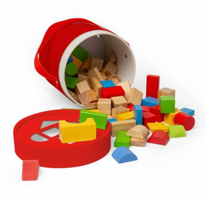 A red children's wooden block storage bin tipped over with colorful wooden blocks spilling out onto a white background. The set includes a red bucket-shaped container, a round lid, and multicolored blocks in natural wood and painted shades of red, yellow, green, blue, and brown.