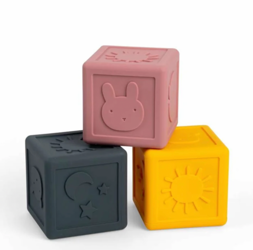 Three silicone baby toy blocks stacked in a pyramid on a white background. The top block is dusty pink with a bunny face, the bottom left is dark gray with a moon and star, and the bottom right is mustard yellow with a sun.