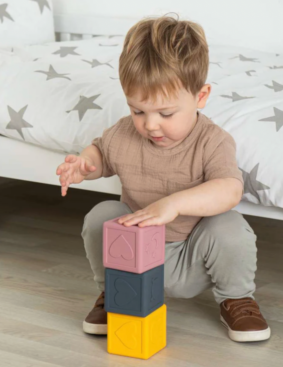 A toddler boy in a brown shirt and grey pants is building a tower of three colorful blocks (pink, dark blue, and yellow) on a light wood floor in front of a bed with grey star-patterned bedding.