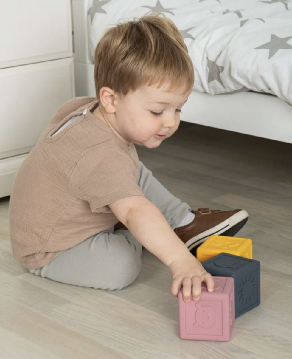 A toddler sits on a light wood floor in a bedroom, reaching for colorful wooden blocks. The child is wearing a beige short-sleeve shirt and light gray pants.