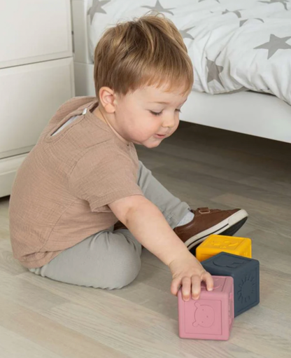 A toddler sits on a light wood floor in a bedroom, reaching for colorful wooden blocks. The child is wearing a beige short-sleeve shirt and light gray pants.