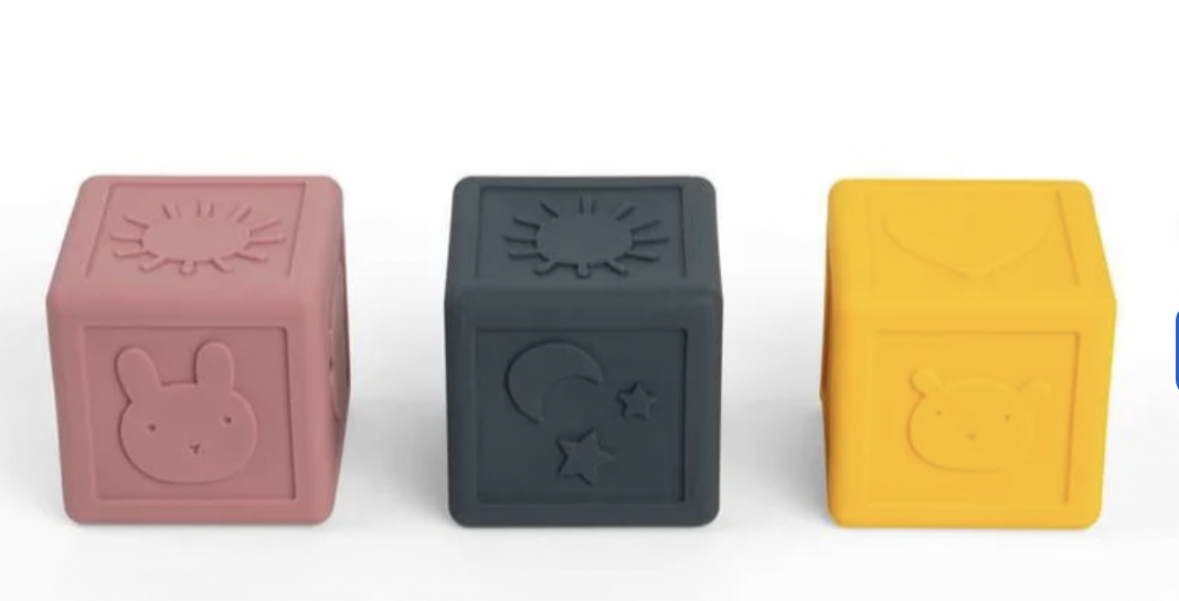 Three silicone baby toy blocks in dusty pink, dark gray, and bright yellow, each embossed with a simple animal or sun motif, arranged on a white background.