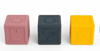 Three silicone baby toy blocks in dusty pink, dark gray, and bright yellow, each embossed with a simple animal or sun motif, arranged on a white background.