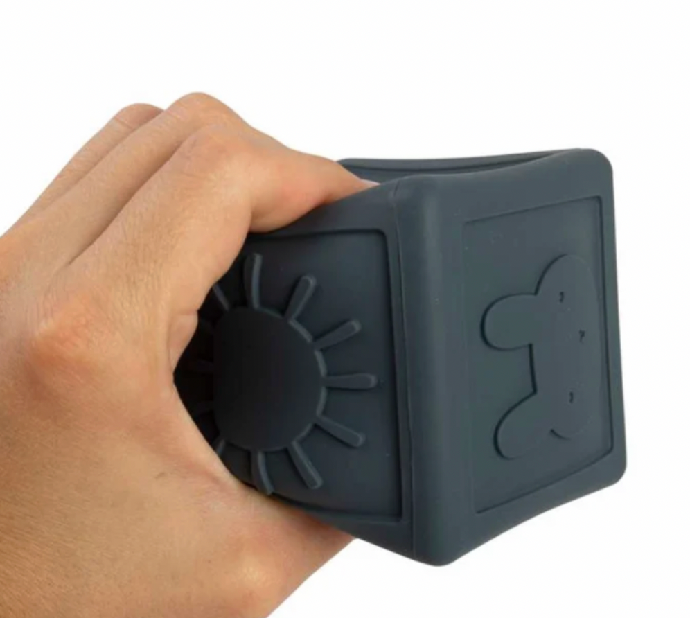 A hand squeezing a dark gray silicone cube toy with embossed animal and sun motifs on a plain white background.