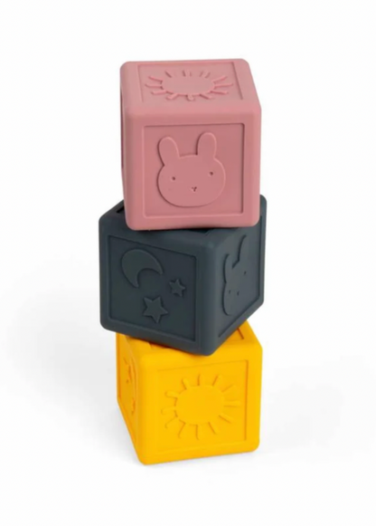 Three stacked silicone baby blocks in dusty pink, dark gray, and mustard yellow, each featuring embossed animal and sun motifs, on a white background.