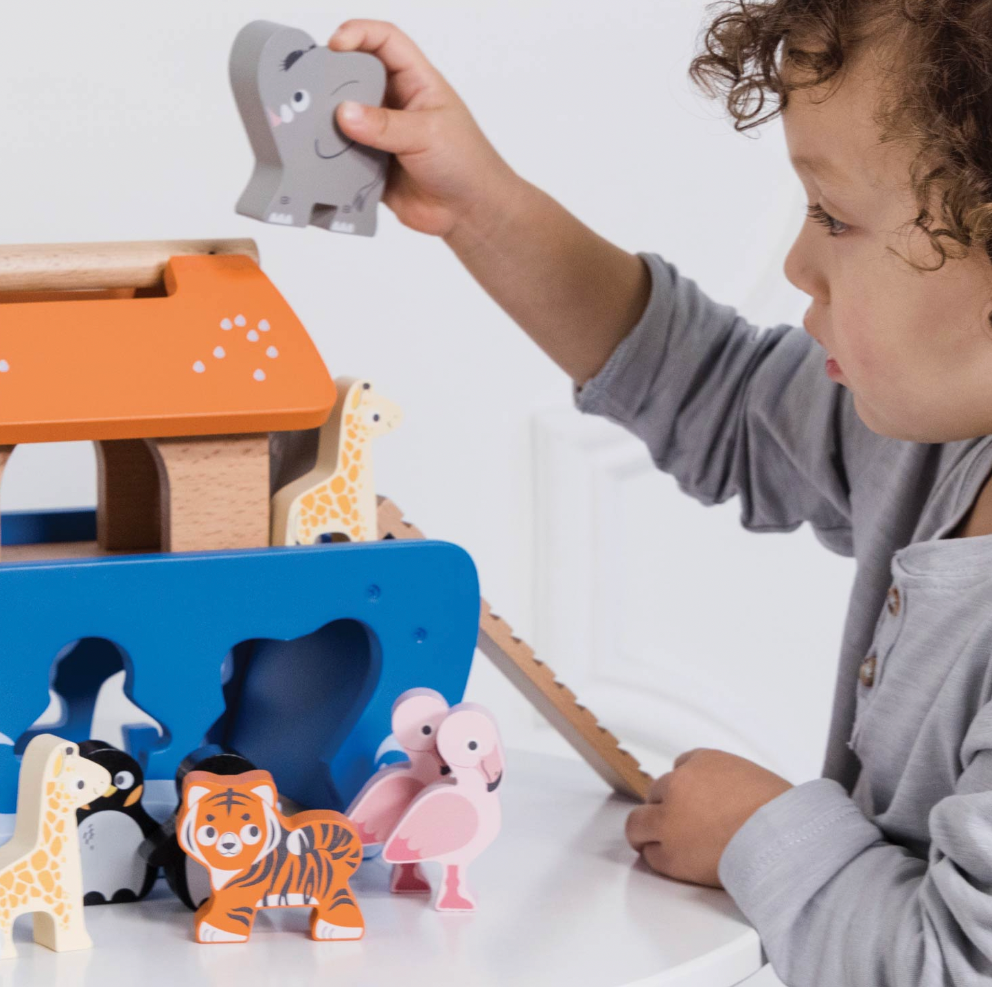 A toddler in a gray long-sleeve shirt plays with a wooden animal sorting and building toy on a white table. The toy includes a blue base with animal cutouts, a wooden house with an orange roof, and several painted animal pieces including a tiger, penguin, giraffe, flamingo, and elephant.