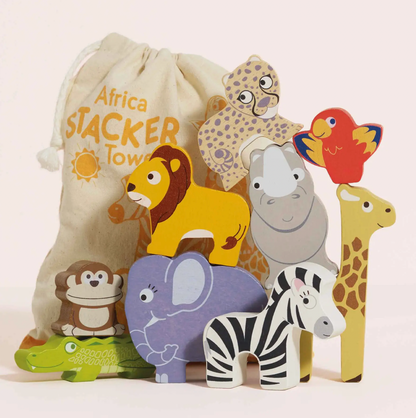 A children's safari-themed stacking toy set with colorful animal cutouts and a cream drawstring bag printed with 'Africa Stacker Town.' The set includes a lion, rhino, elephant, zebra, giraffe, crocodile, monkey, leopard, and bird, arranged in front of the bag on a light pink background.