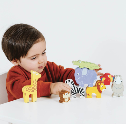 A young child wearing a rust-red sweatshirt sits at a white table, playing with a set of colorful wooden animal toys including a giraffe, zebra, lion, elephant, crocodile, monkey, parrot, and sheep.