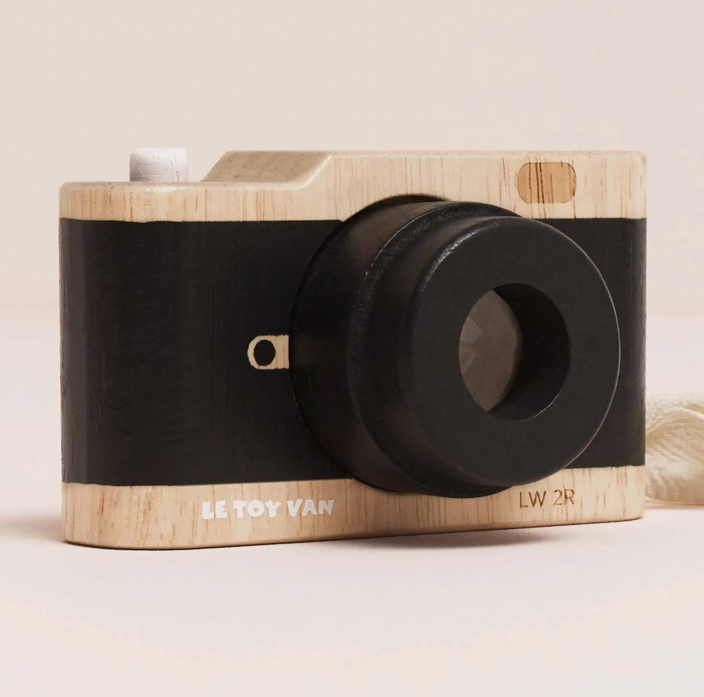 A toy camera with a light natural wood body and a wide black band around the middle, photographed on a plain light beige background. The front shows a large black lens, a small white button, a tiny gold-toned detail, and printed text reading 'LE TOY VAN' and 'LW 2R'.