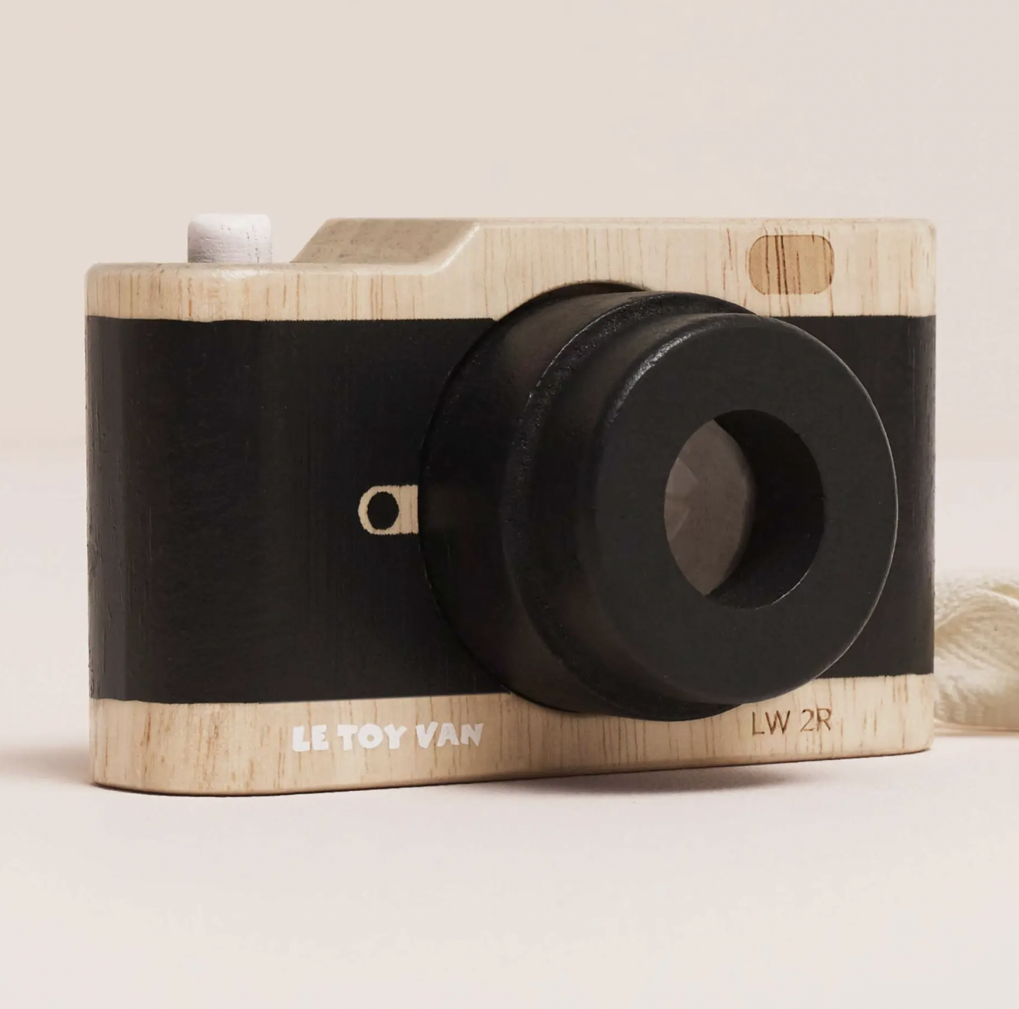 A toy camera with a light natural wood body and a wide black band around the middle, photographed on a plain light beige background. The front shows a large black lens, a small white button, a tiny gold-toned detail, and printed text reading 'LE TOY VAN' and 'LW 2R'.
