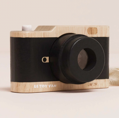 A toy camera with a light natural wood body and a wide black band around the middle, photographed on a plain light beige background. The front shows a large black lens, a small white button, a tiny gold-toned detail, and printed text reading 'LE TOY VAN' and 'LW 2R'.