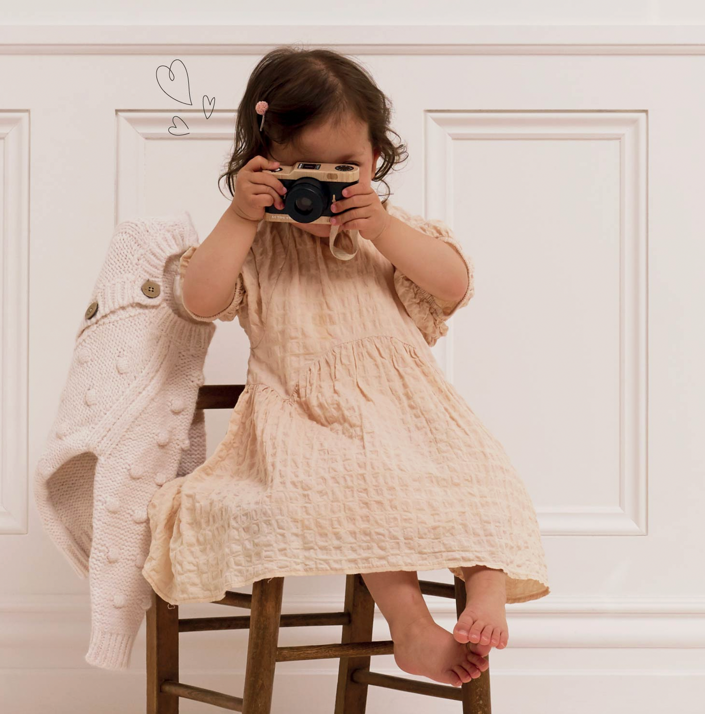 A toddler girl sits on a wooden stool in front of a white paneled wall, holding a retro camera to her face. She is wearing a textured cream-colored cotton dress, and a cream knit cardigan is draped over the stool beside her.