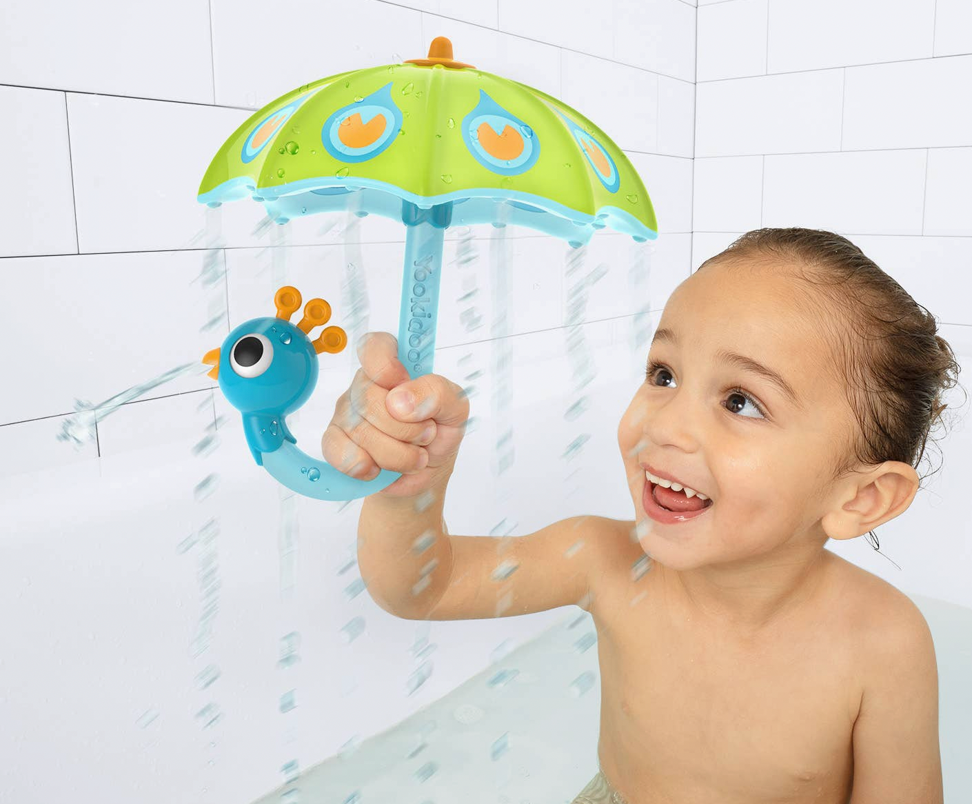 A smiling young child holding a colorful toy shower umbrella in a bathtub, with water spraying down. The toy has a green umbrella top with orange and blue details and a blue caterpillar-shaped handle.