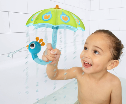 A smiling young child holding a colorful toy shower umbrella in a bathtub, with water spraying down. The toy has a green umbrella top with orange and blue details and a blue caterpillar-shaped handle.