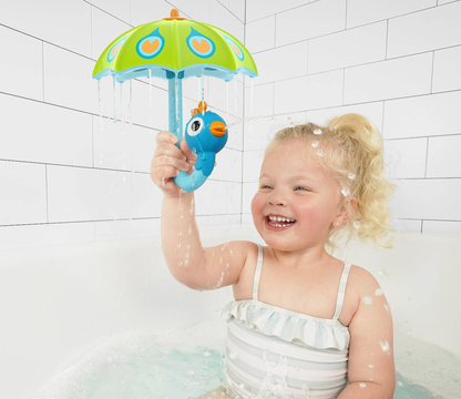 A young child with blonde hair sitting in a white bathtub, holding a blue rubber duck toy under a green and blue toy umbrella that is spraying water.