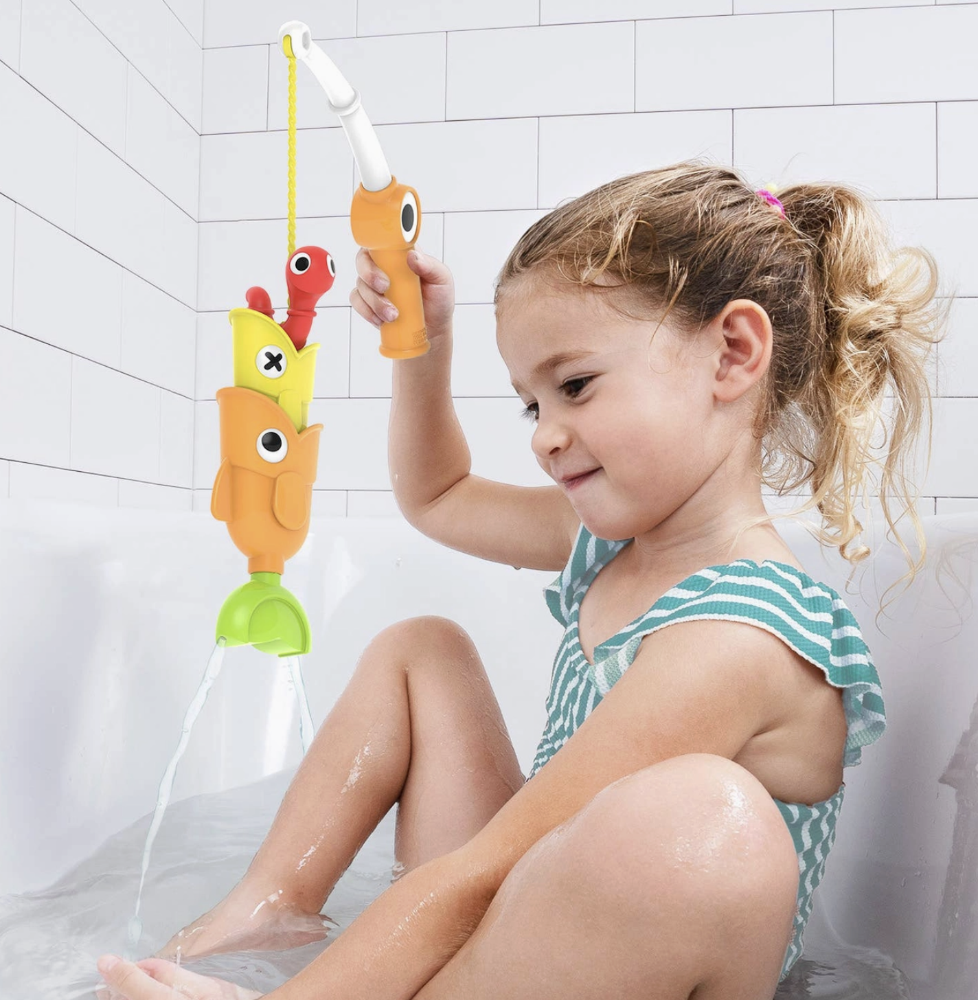 A young girl sitting in a bathtub plays with a colorful fish-shaped bath toy held by a handheld shower attachment. The toy features orange, yellow, red, and green fish shapes with cartoon eyes.