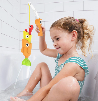A young girl sitting in a bathtub plays with a colorful fish-shaped bath toy held by a handheld shower attachment. The toy features orange, yellow, red, and green fish shapes with cartoon eyes.