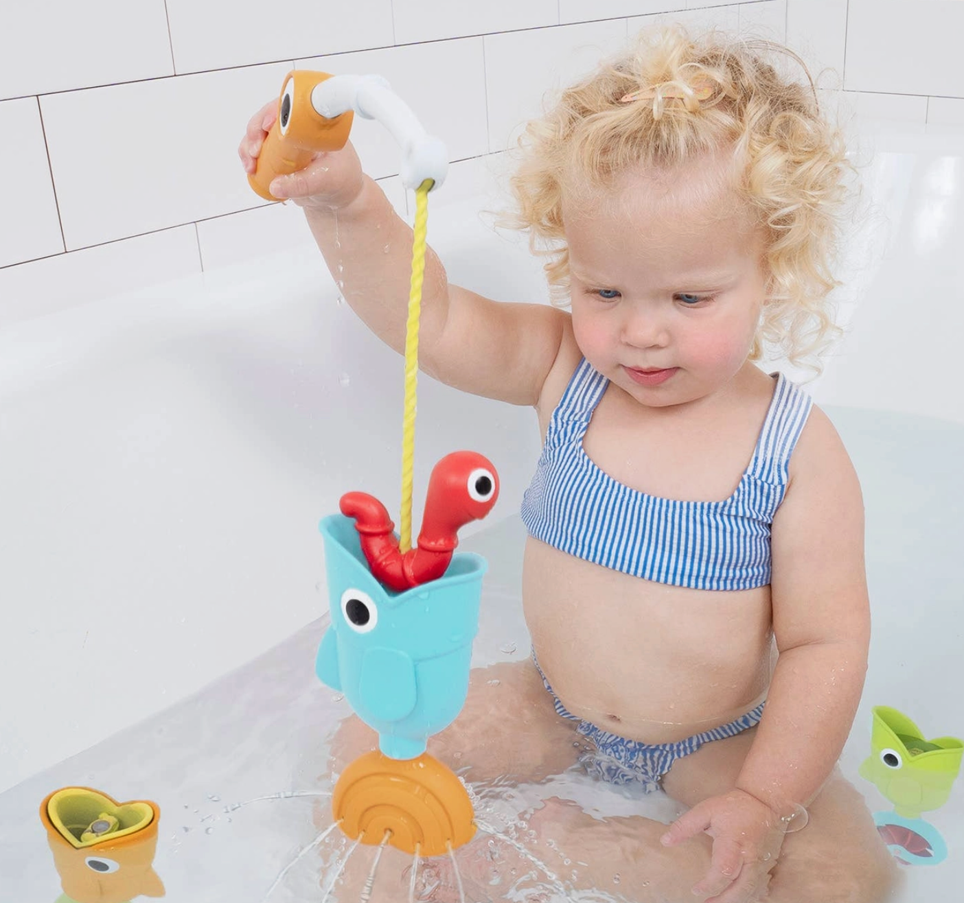 A toddler with curly blonde hair sits in a white bathtub, playing with a colorful toy fishing set. The child is wearing a blue and white striped swimsuit and holds a toy fish above a light blue bucket with a red worm and a yellow rope.