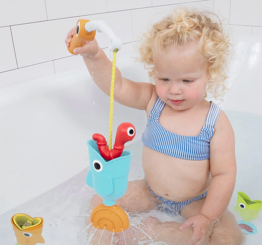 A toddler with curly blonde hair sits in a white bathtub, playing with a colorful toy fishing set. The child is wearing a blue and white striped swimsuit and holds a toy fish above a light blue bucket with a red worm and a yellow rope.