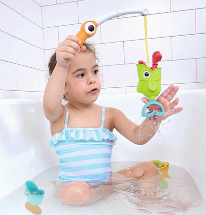 A young child sitting in a white bathtub with water, holding a colorful toy fishing rod with a green frog-shaped hook and blue ring. The child is wearing a light blue and white striped swimsuit.